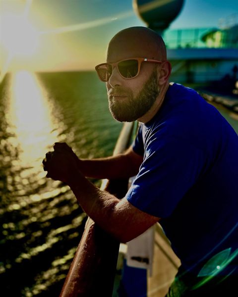 Brandon Sainsbury, founder of Green Country Front Desk, leaning on a rail
                      at sunset with the water behind him.
